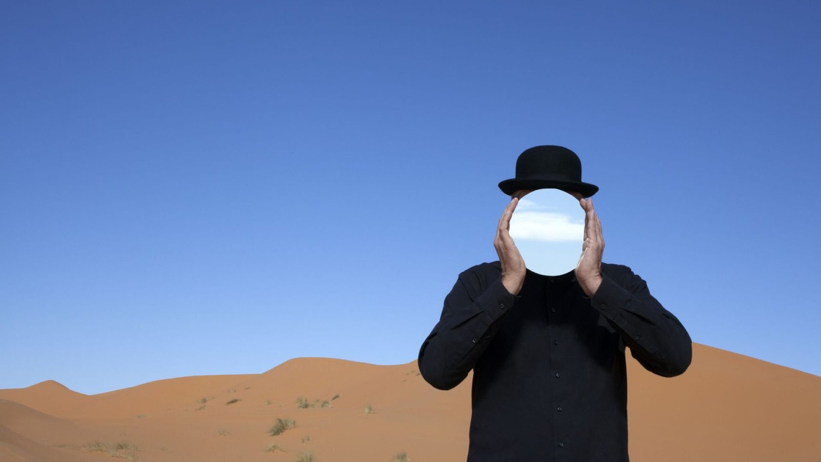Desierto Morocco, Merzouga, Erg Chebbi, man wearing a bowler hat holding mirror in front of his face in deser